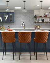 Three modern bar stools with chestnut brown quilted leather upholstery and sleek black metal legs, situated at a kitchen island.