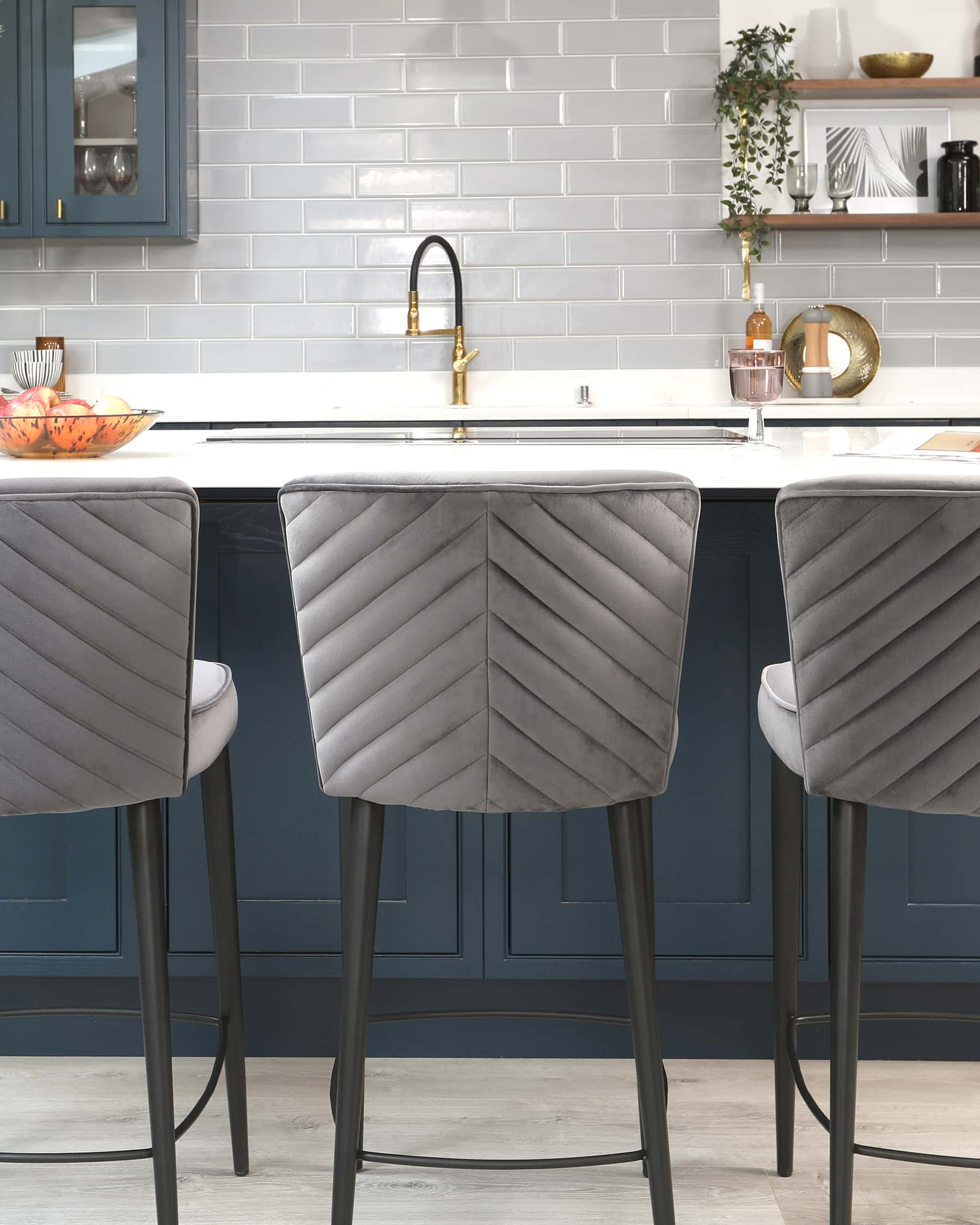 Three contemporary bar stools with grey upholstered seats and chevron-pattern stitching. The stools feature slender black metal legs with footrests.