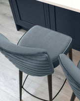 Modern grey upholstered bar stool with a horizontal channel-tufted backrest and cushioned seat, featuring slender black metal legs with a footrest.