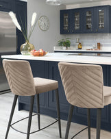 Elegant modern bar stools with taupe upholstered seats and chevron patterned backrests, featuring slender black metal legs with footrests.