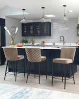 Three modern upholstered bar stools with a herringbone pattern and neutral taupe fabric seat, elegant curved backrest, and sleek black metal legs, positioned at a white kitchen island with a marble countertop.