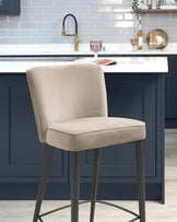 Elegant modern bar stool with a beige upholstered seat and backrest on a sleek black metal frame, showcased in a kitchen with dark blue cabinetry and white marble countertop.