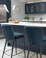 Two elegant blue upholstered bar stools with chevron stitching and slim metal legs are positioned at a white kitchen island with a modern design.