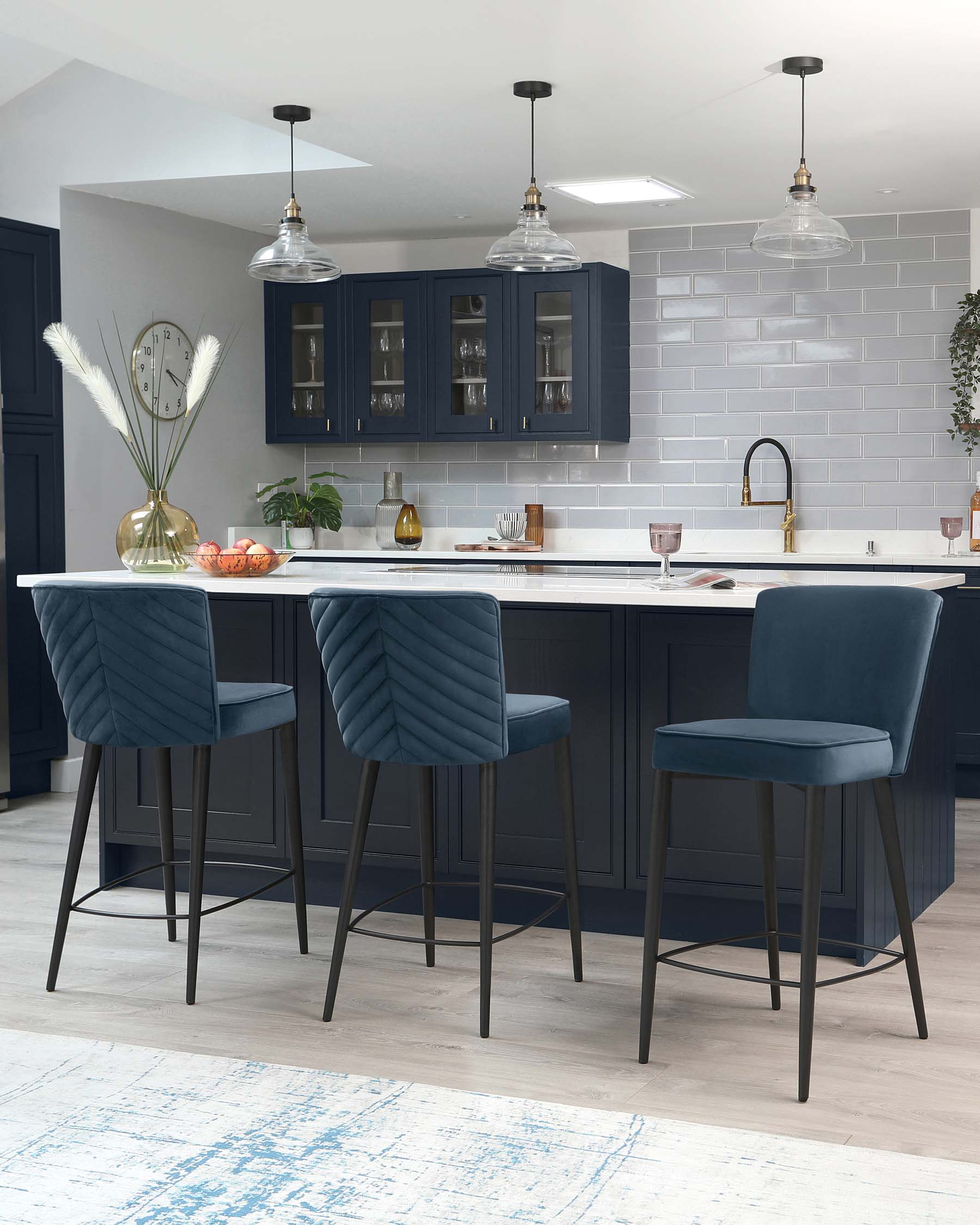 Three contemporary navy blue barstools with channel tufting on the backrest and smooth upholstered seating, paired with slim black metal legs that include footrests, arranged at a modern kitchen island.