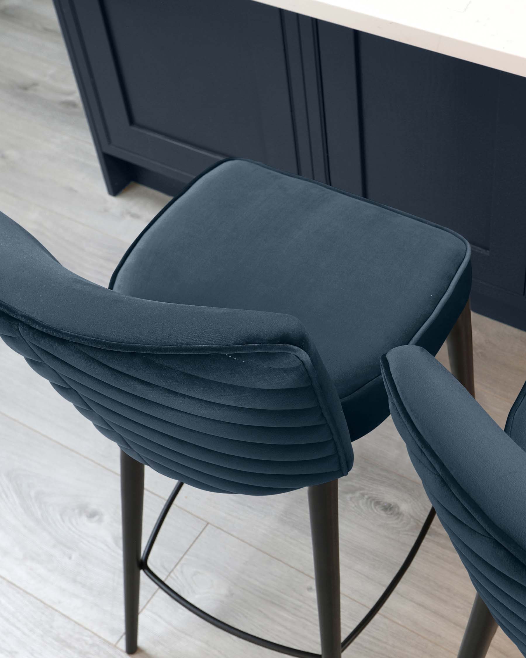 Modern dark blue upholstered dining chair with distinctive horizontal channel stitching on the backrest, a comfortably padded seat, and slender metal legs in a matte finish, set against a kitchen island with dark cabinetry.