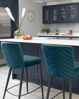 Two modern teal velvet bar stools with chevron patterned backrests and matte grey metal legs, showcased in a contemporary kitchen with dark blue cabinetry and white countertops.