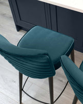 A modern stool with dark teal upholstery and horizontal channel stitching. The stool features a comfortable backrest and seat cushion on an elegant black metal frame with slender legs and a footrest. The design offers a chic, contemporary look suitable for a kitchen or bar setting.