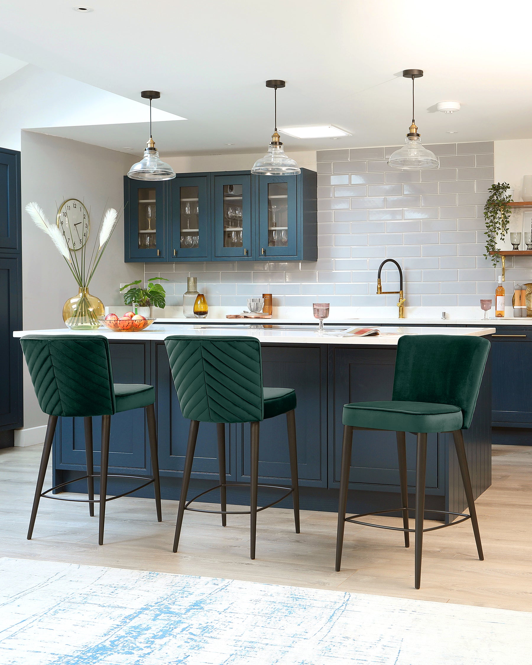 Modern kitchen interior showcasing a set of three luxurious green velvet bar stools with channel tufting and black metal legs, aligned at a sleek kitchen island with a white marble countertop.