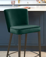 Elegant emerald green velvet bar stool with a high backrest and a cushioned seat, mounted on a sleek metal frame with four slender legs and footrests. The luxurious colour and texture contrast with the contemporary kitchen setting in the background.