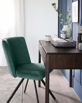 Elegant home office setup featuring a dark wooden writing desk with sleek, straight lines and a modern green velvet chair with black metal legs. Desk accessories include a laptop, notepad, and decorative vase with greenery.