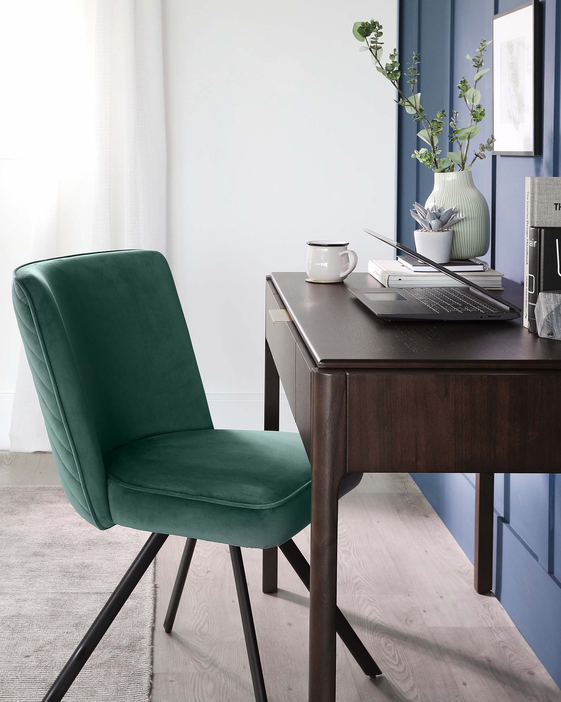 Elegant home office setup featuring a dark wooden writing desk with sleek, straight lines and a modern green velvet chair with black metal legs. Desk accessories include a laptop, notepad, and decorative vase with greenery.