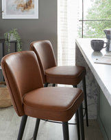 Two modern counter-height stools with caramel brown faux leather upholstery and black metal legs, situated at a white marble countertop.