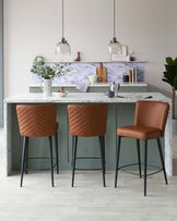 Three modern bar stools with caramel brown faux leather upholstery and quilted backrests, paired with slim black metal legs including footrests. These stools are positioned in front of a kitchen island with a white marble countertop and matte green cabinetry.