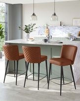 Elegant modern kitchen interior featuring a set of three quilted caramel brown leather bar stools with slender black metal legs perched at a white marble breakfast bar.