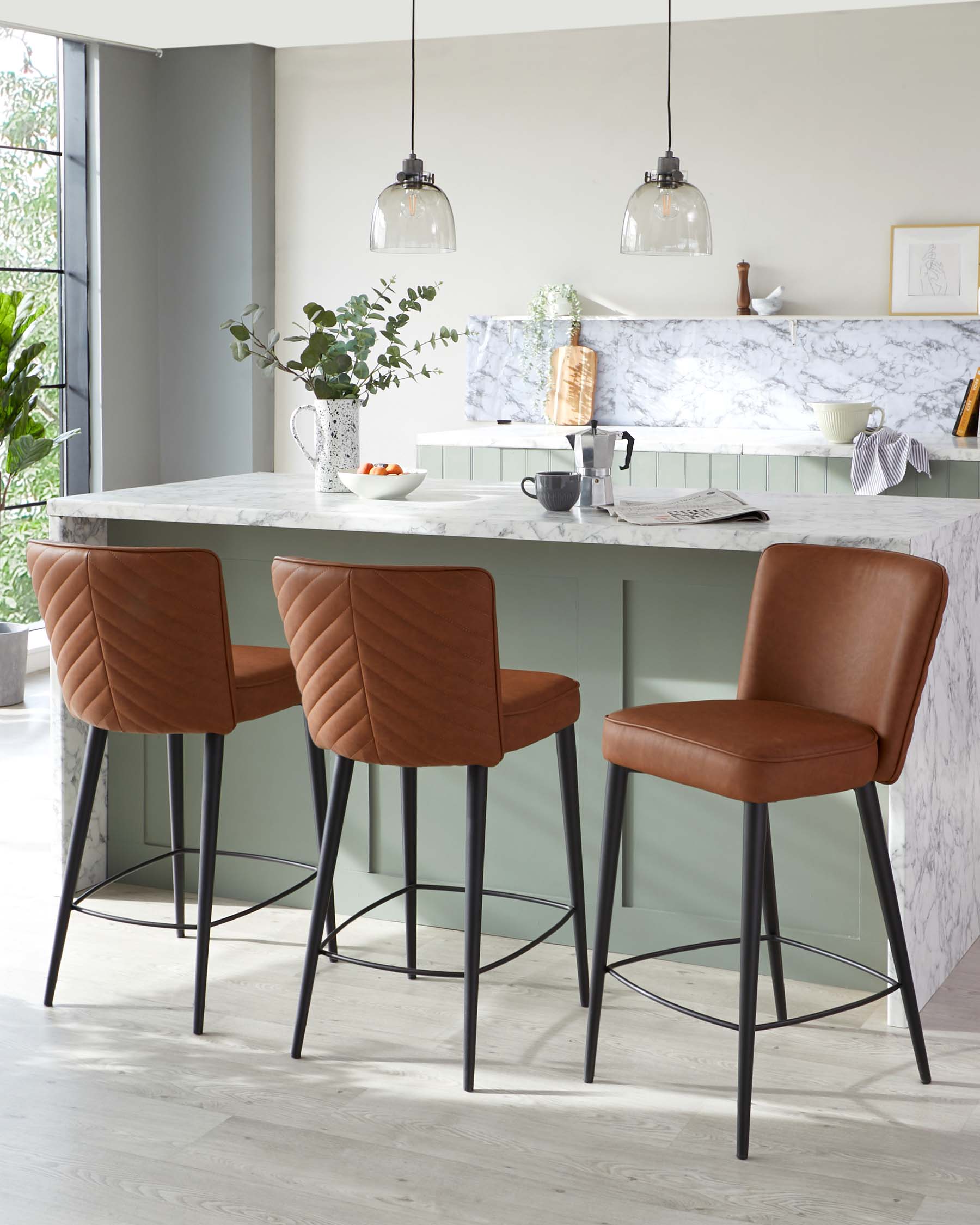 Elegant modern kitchen interior featuring a set of three quilted caramel brown leather bar stools with slender black metal legs perched at a white marble breakfast bar.