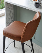 Modern bar stool with a luxurious tan faux leather seat and a curved, quilted backrest. The stool features a minimalist black metal frame with sleek, straight legs and a circular footrest. The design is contemporary, with an emphasis on comfort and clean lines, ideal for modern kitchen or bar settings.