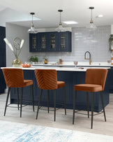 Three contemporary bar stools with sleek metal legs and terracotta-hued upholstered seats and backs featuring a chevron pattern. The stools are positioned at a kitchen island with a white countertop.