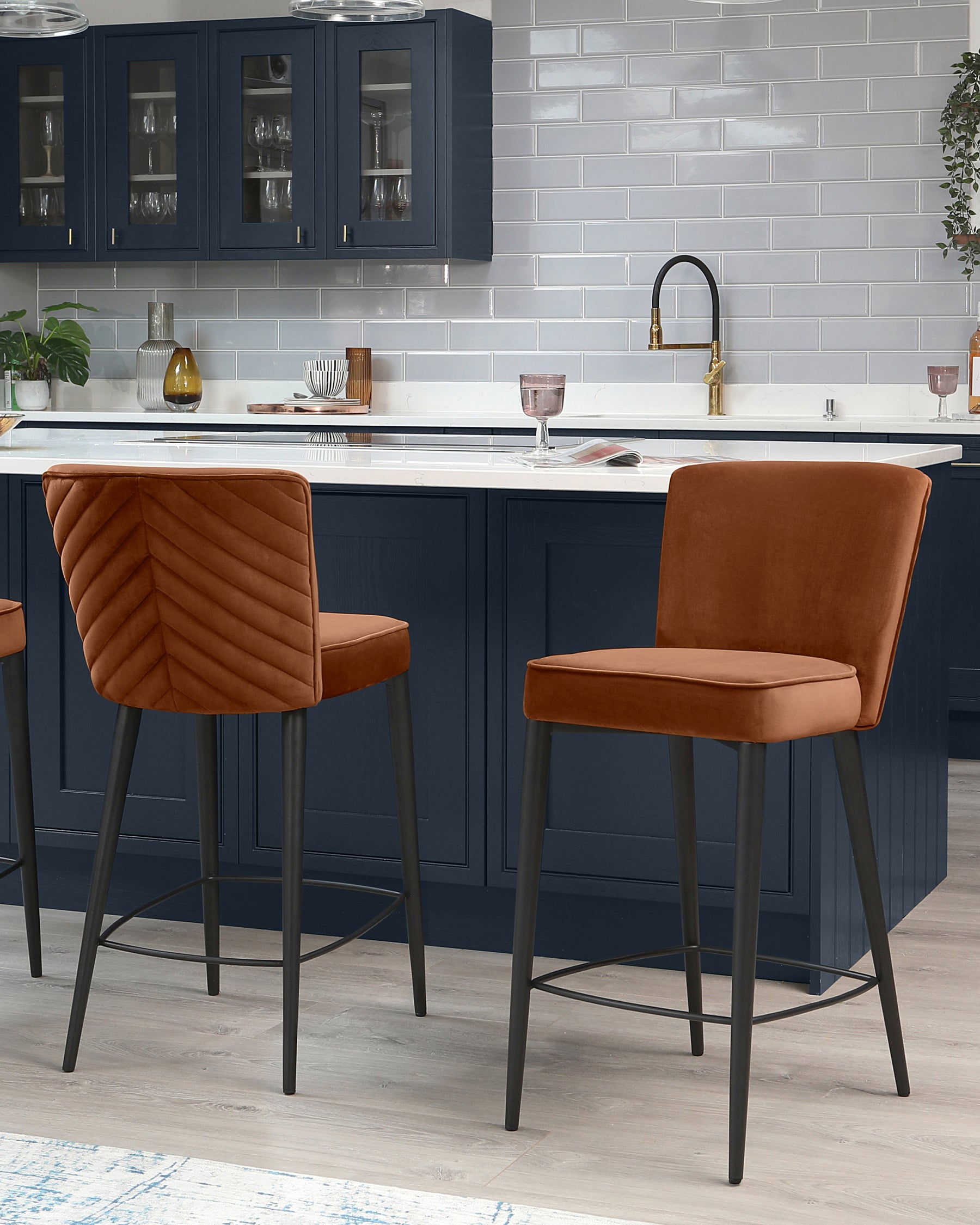 Two modern, burnt orange upholstered bar stools with a channel tufted backrest and sleek black metal legs positioned at a kitchen island.