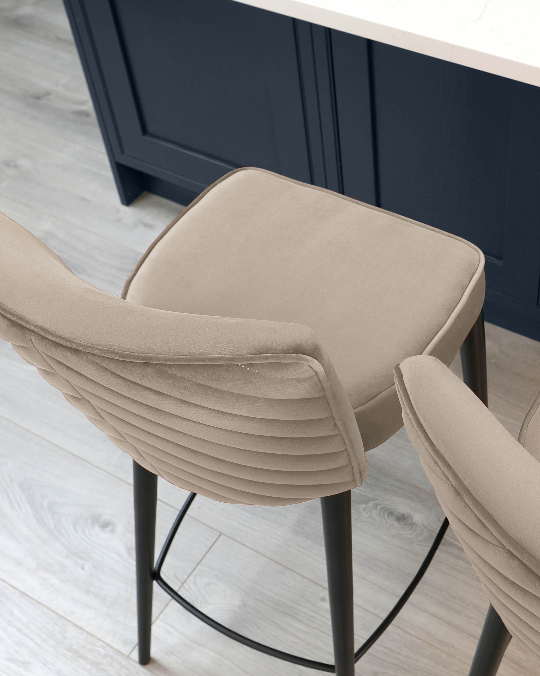 Modern beige upholstered bar stool with channel tufting on the backrest and a sleek black metal frame, positioned against a navy blue kitchen island with white countertop, over light wooden flooring.