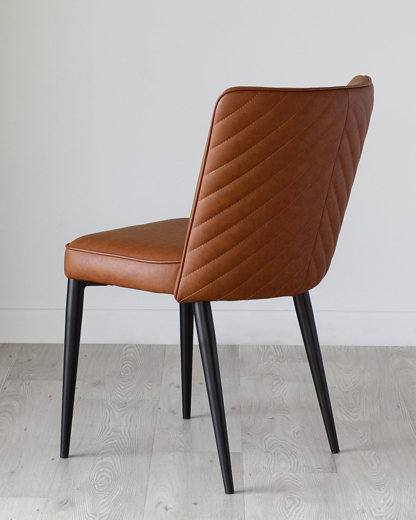 Elegant modern dining chair with a curved, padded backrest featuring quilted diamond stitching, upholstered in a rich caramel brown faux leather, complemented by four sleek, black tapered metal legs.