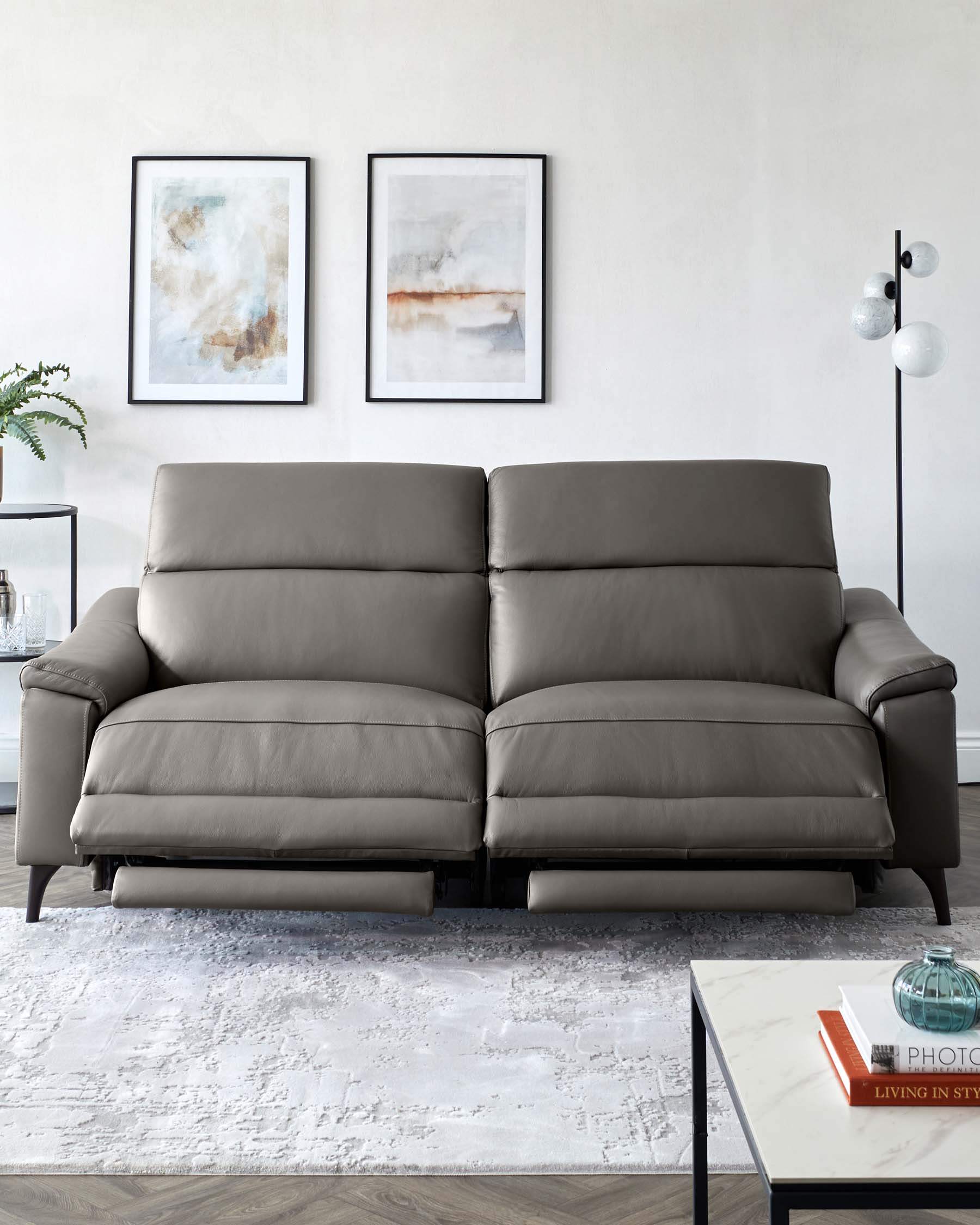 Modern grey leather sofa with plush cushions and sleek armrests, accompanied by a white marble-top coffee table with black metal frame, set on a textured off-white area rug. An elegant floor lamp with spherical shades and abstract wall art complete the contemporary living space.