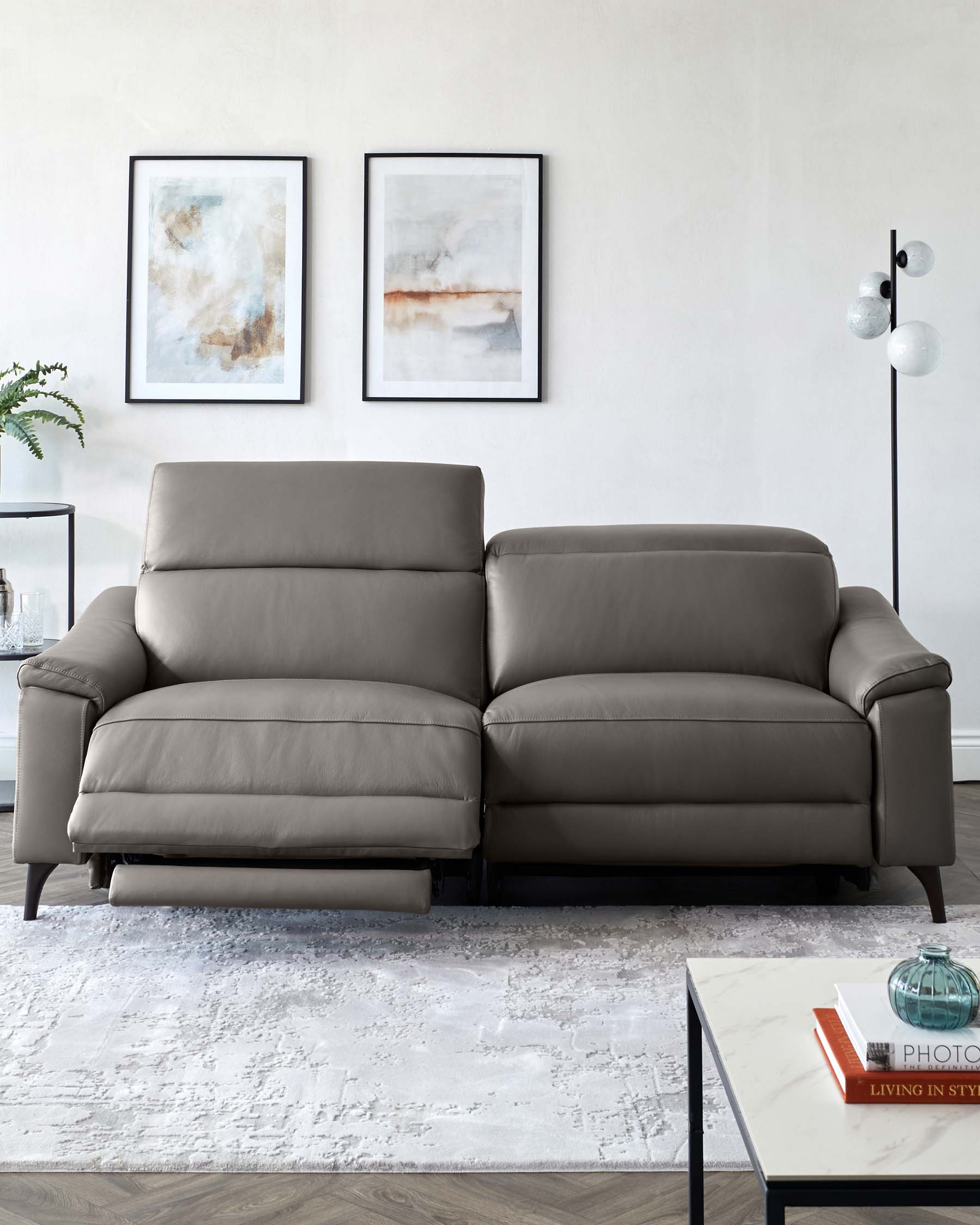 Elegant modern grey leather sofa with plush cushioning and a streamlined design, featuring wide armrests and dark wooden legs. A minimalist white marble-topped coffee table with a black metal frame sits in the foreground, and a sleek black floor lamp with spherical white glass shades stands to the right. The setting is complemented by a textured off-white area rug and abstract wall art in neutral tones.