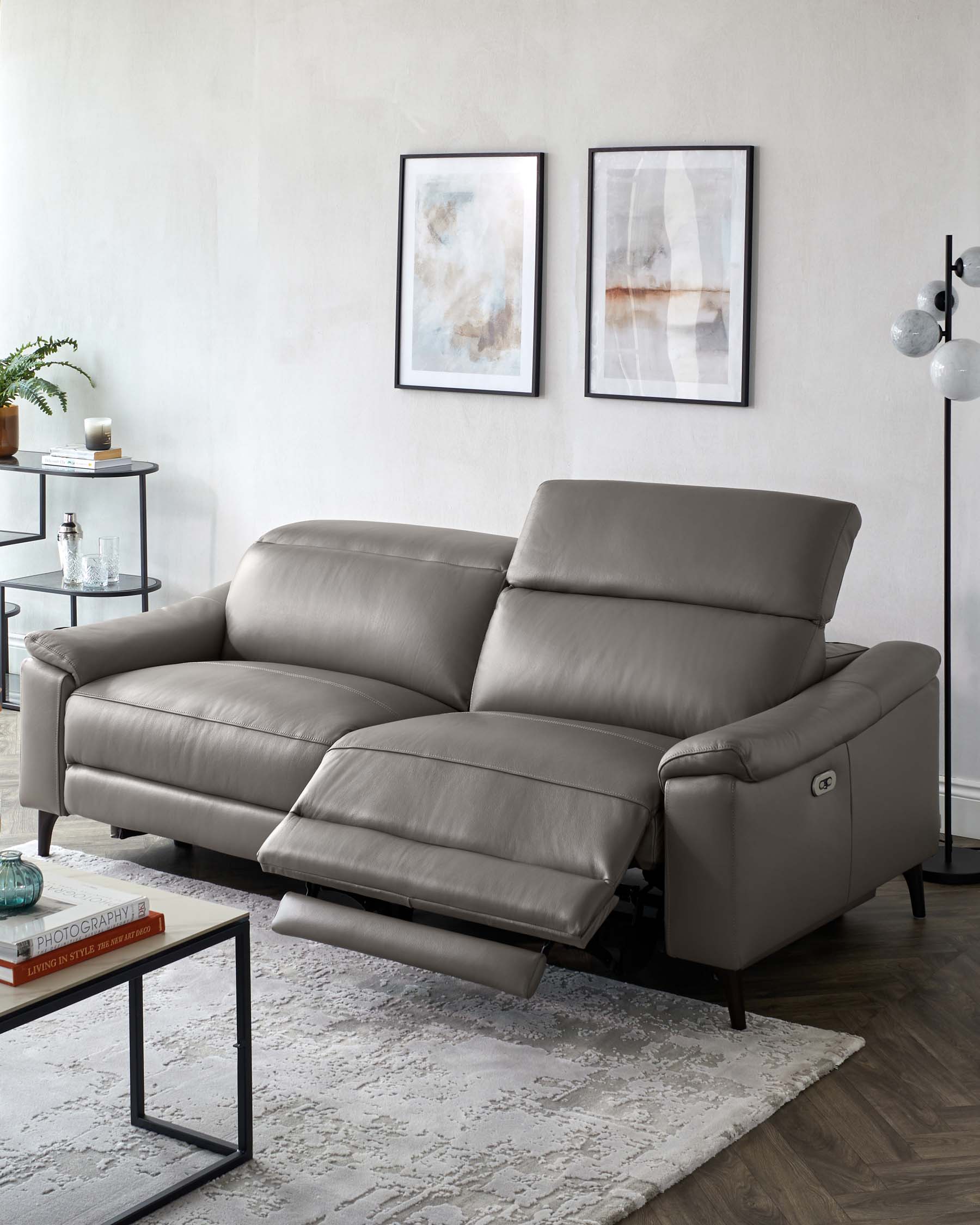 A modern grey leather reclining sofa with a chaise on the left side and a reclined footrest on the right, complemented by a minimalist glass-top end table with a black metal frame on the side. The sofa is adorned with subtle stitching details and pillow-top armrests, set upon a textured off-white area rug over dark hardwood flooring.