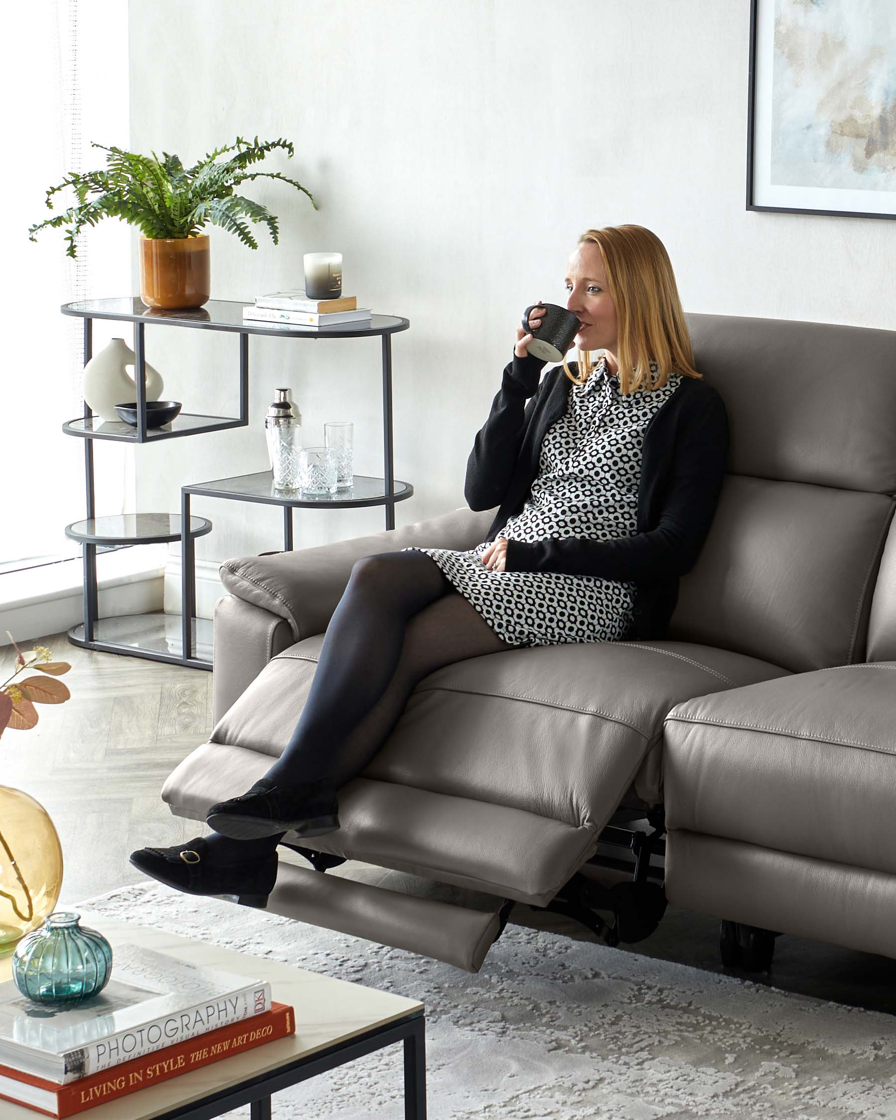 A modern living room furnished with a sleek light grey leather sectional sofa featuring an extended footrest, complemented by a trendy matte black tiered circular side table with layers for display and storage. In the foreground, a low-profile rectangular coffee table with a glossy white surface and black metal frame that exhibits a collection of decorative books and a small glass vase. The room has a cosy yet sophisticated ambiance, ideal for lounging or casual entertaining.