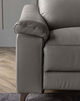 A close-up of a contemporary light grey leather sofa showcasing its plush armrest, detailed stitching, and cushioned backrest. The elegant furniture piece rests on a dark hardwood floor against a soft grey wall, suggesting a modern and sleek design suitable for a variety of home interiors.