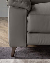 Close-up of a section of a modern grey leather sofa with plush cushioning and detailed stitching, set on a two-tone grey and beige textured rug over wooden flooring. The sofa is elevated on a dark wooden tapered leg.