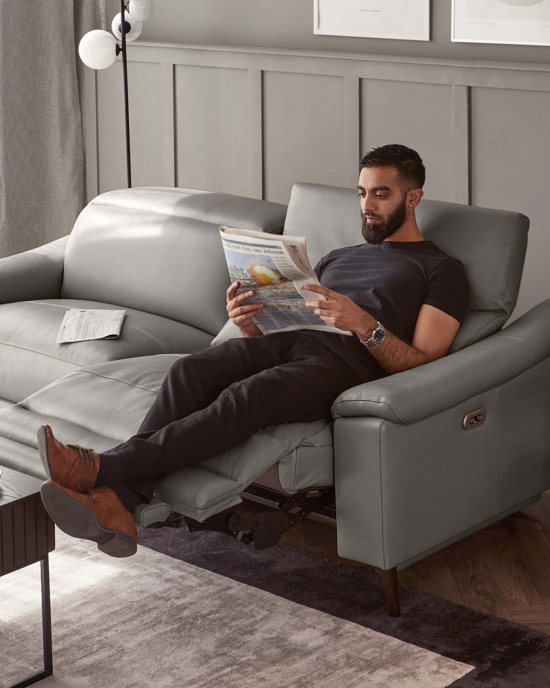 A modern grey leather recliner with a sleek design and dark wooden legs, featuring plush cushioning and an adjustable footrest extended, set in a contemporary room with a matching grey wall panelling.