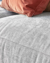 Close-up view of a textured grey fabric, possibly an upholstered piece of furniture, with a dusty rose-coloured pillow with braided trim detail resting on it.