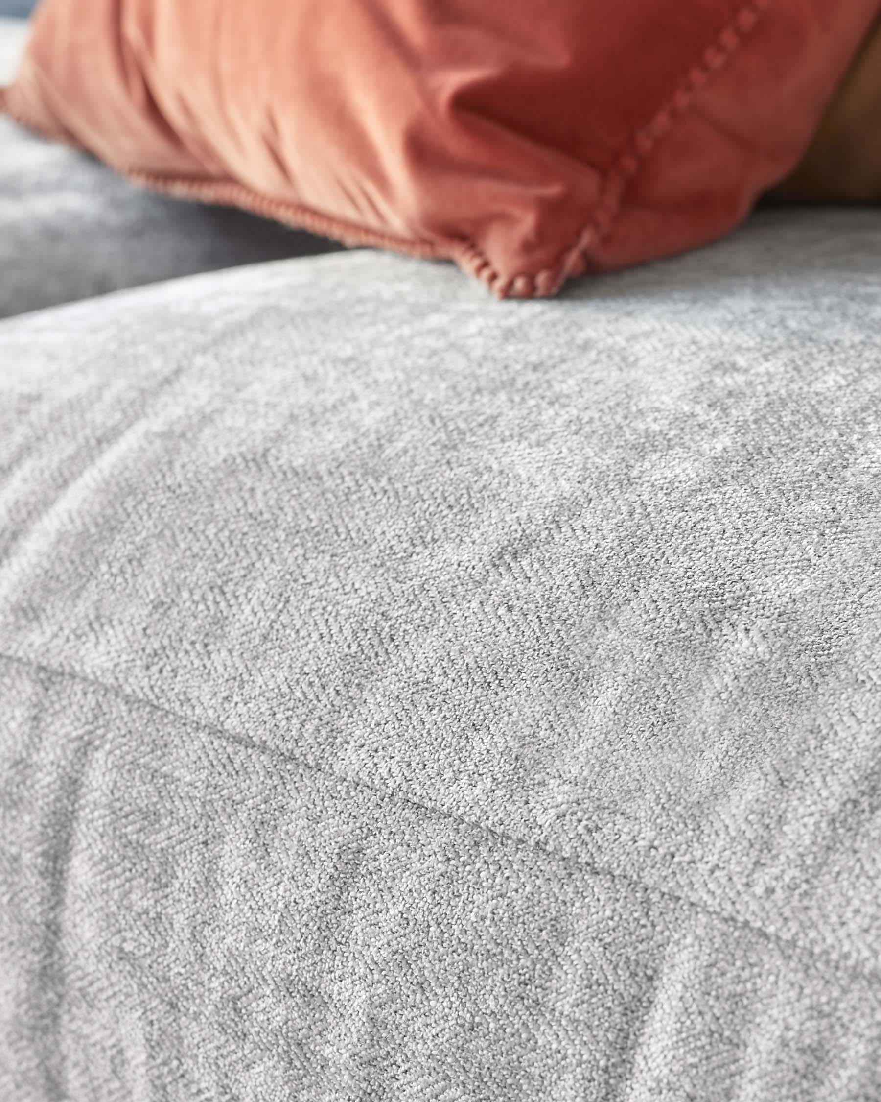 Close-up view of a textured grey fabric sofa with a rust-coloured throw pillow, showcasing the detailed stitching and soft fabric.