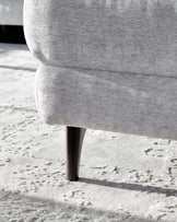 Close-up of a light grey upholstered chair with a textured fabric and a tapered dark wooden leg, set on a white patterned area rug.