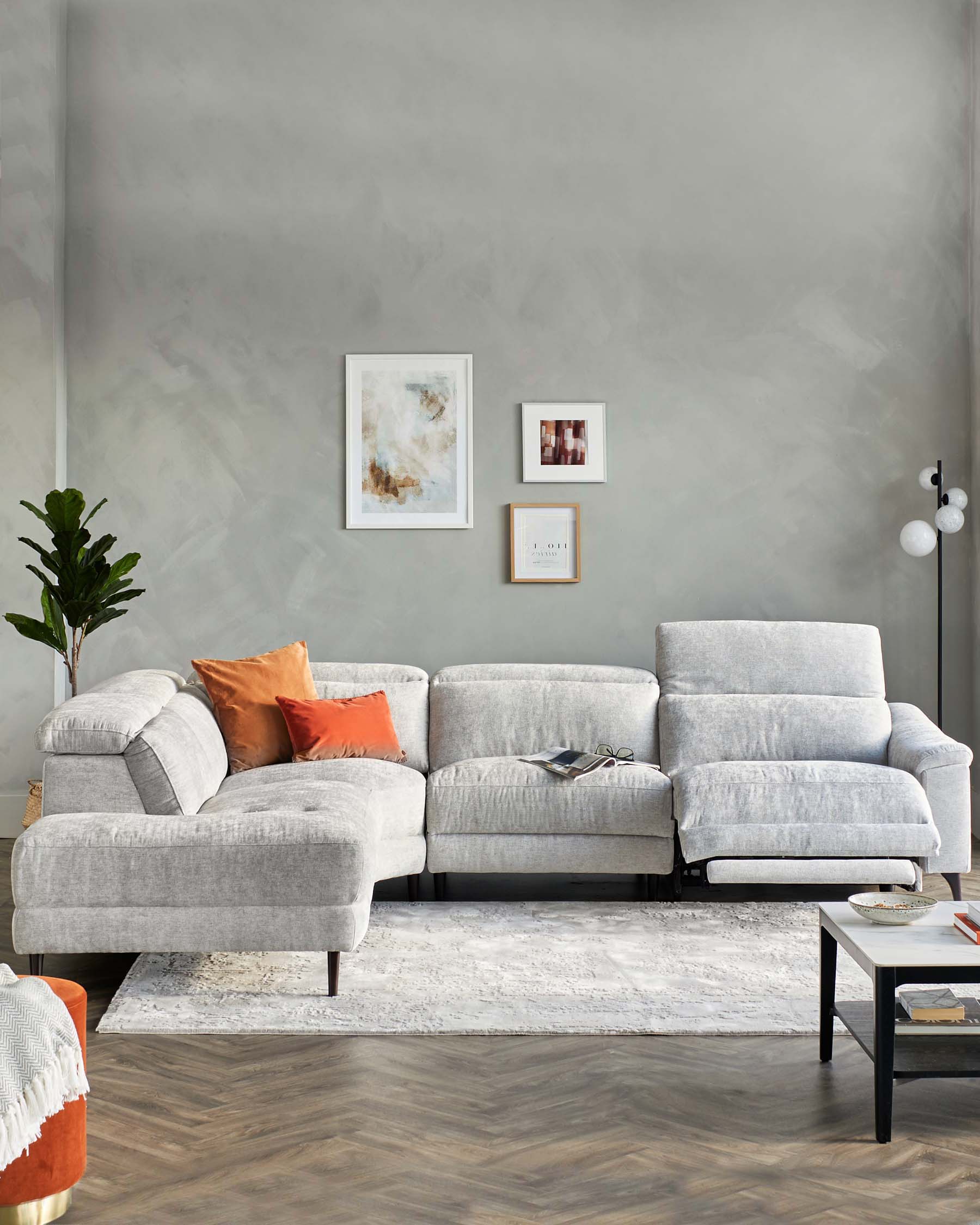 Modern light grey sectional sofa with chaise on the left and recliner features, accessorized with orange and white throw pillows. In front, a rectangular white and grey area rug lays on a wooden floor. To the right, a minimalistic black coffee table holding books and a decorative bowl.