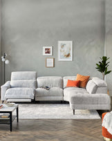 Light grey fabric sectional sofa with plush cushions and a chaise on the left side, paired with a small rectangular black coffee table with a white top, resting on a textured white area rug. A modern black floor lamp stands to the left. Decor includes wall art, throw pillows in orange and neutral tones, and a potted plant to the right, creating an inviting and stylish living space.