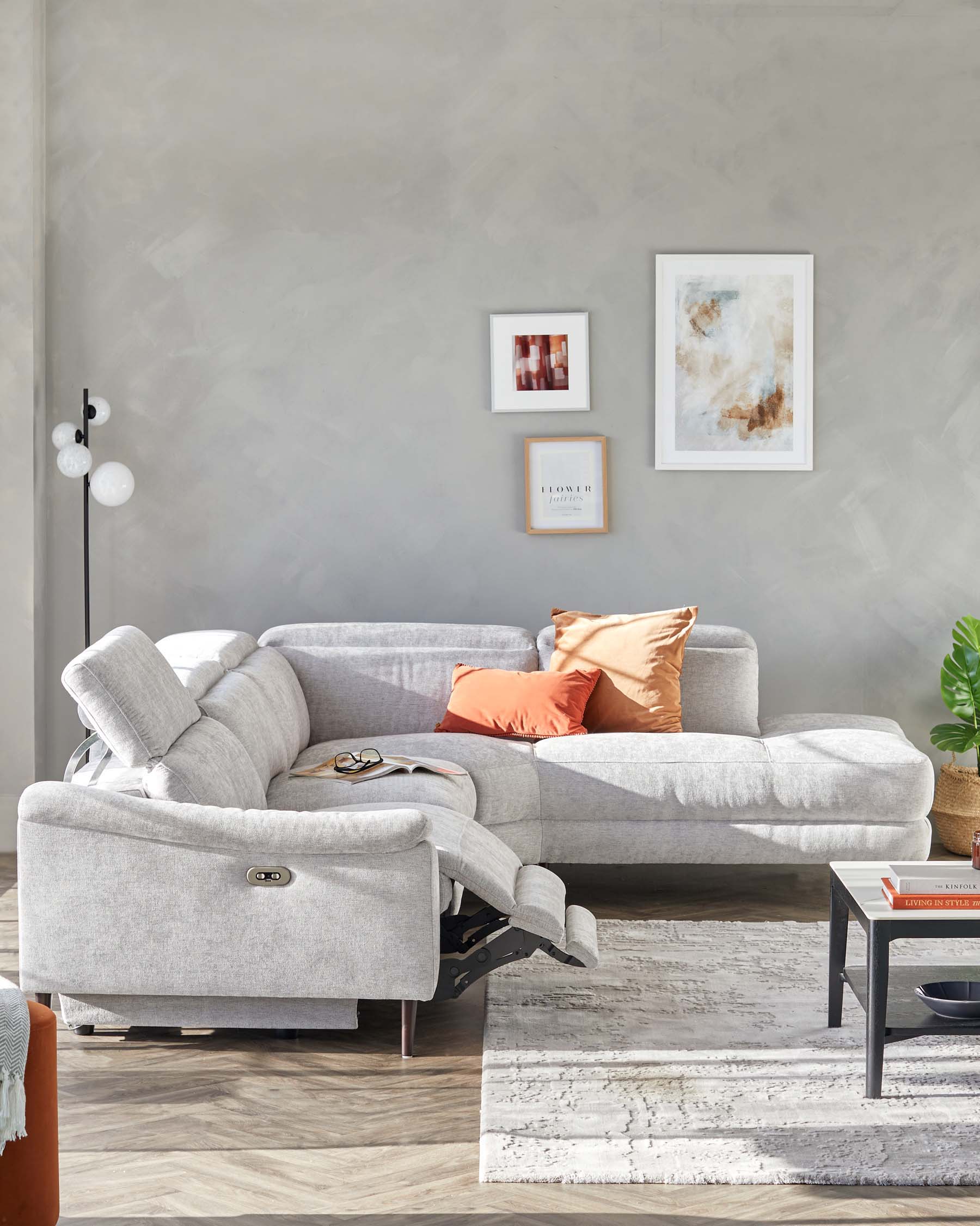 A contemporary light grey fabric sectional sofa with a reclining end, complemented by three decorative cushions in shades of orange. In front of it lies a distressed white and grey area rug. To the right stands a small black end table with a contrasting orange tabletop book, beside a potted green plant.