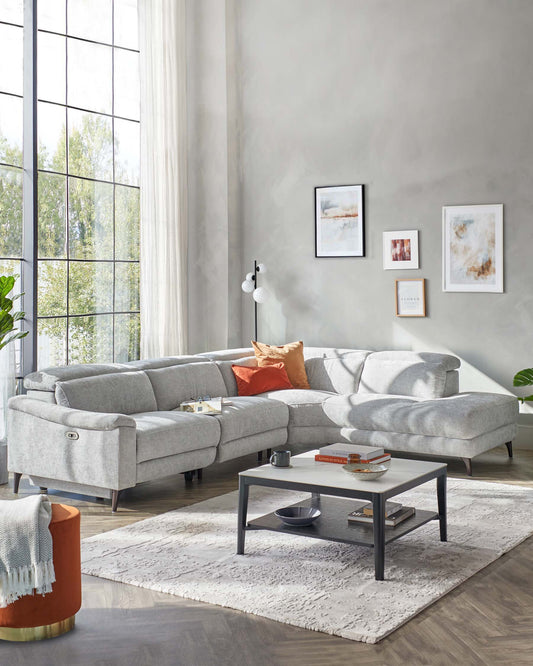 Modern light grey sectional sofa with chaise on silver metal legs, small round orange side table with golden base, rectangular black and grey coffee table with a lower shelf, and a white textured area rug.