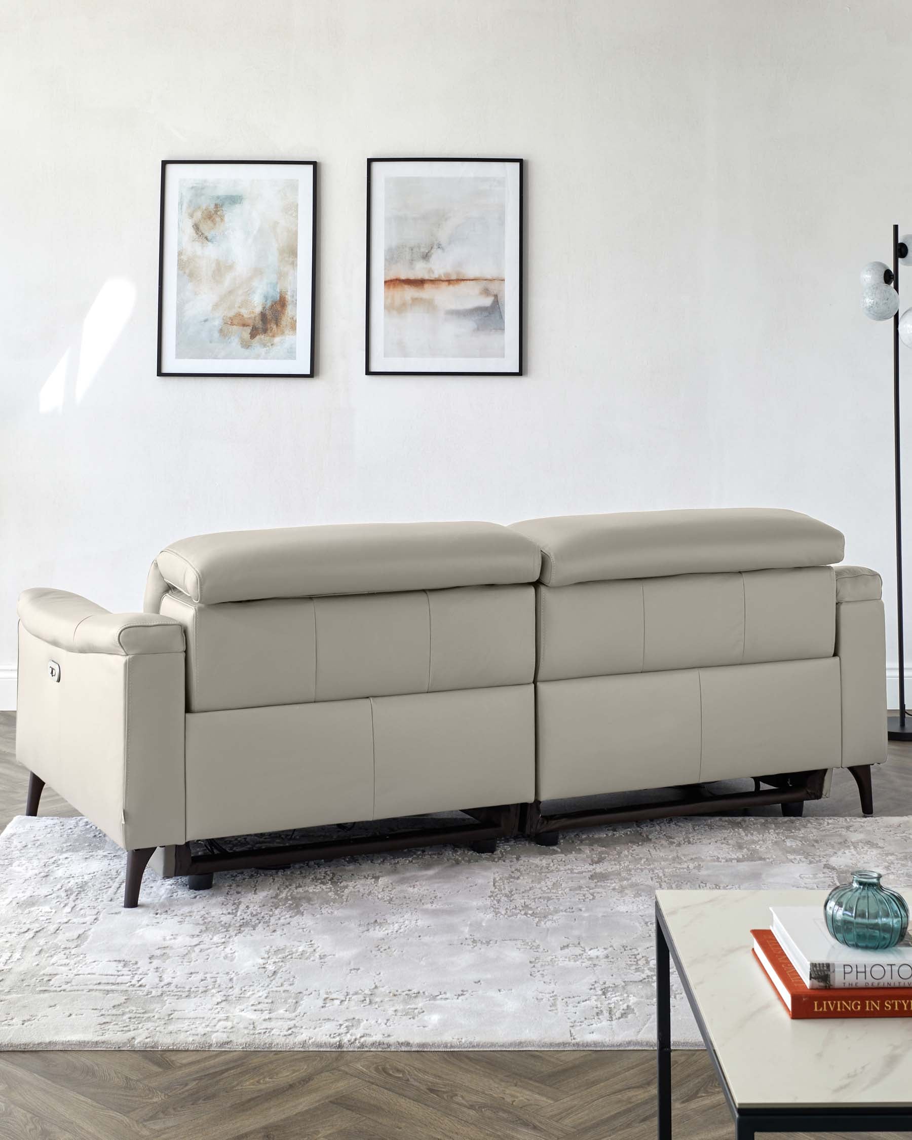 Elegant light grey leather sectional sofa featuring a streamlined design with a right-facing chaise. The couch is accented by dark wooden legs and is staged on a textured white area rug over a hardwood floor. A modern glass-top coffee table with a black metal frame sits in the foreground, complemented by a teal glass vase and books. Two abstract framed art pieces hang on the wall above the sofa, and there is a contemporary floor lamp with a glass globe on the right.