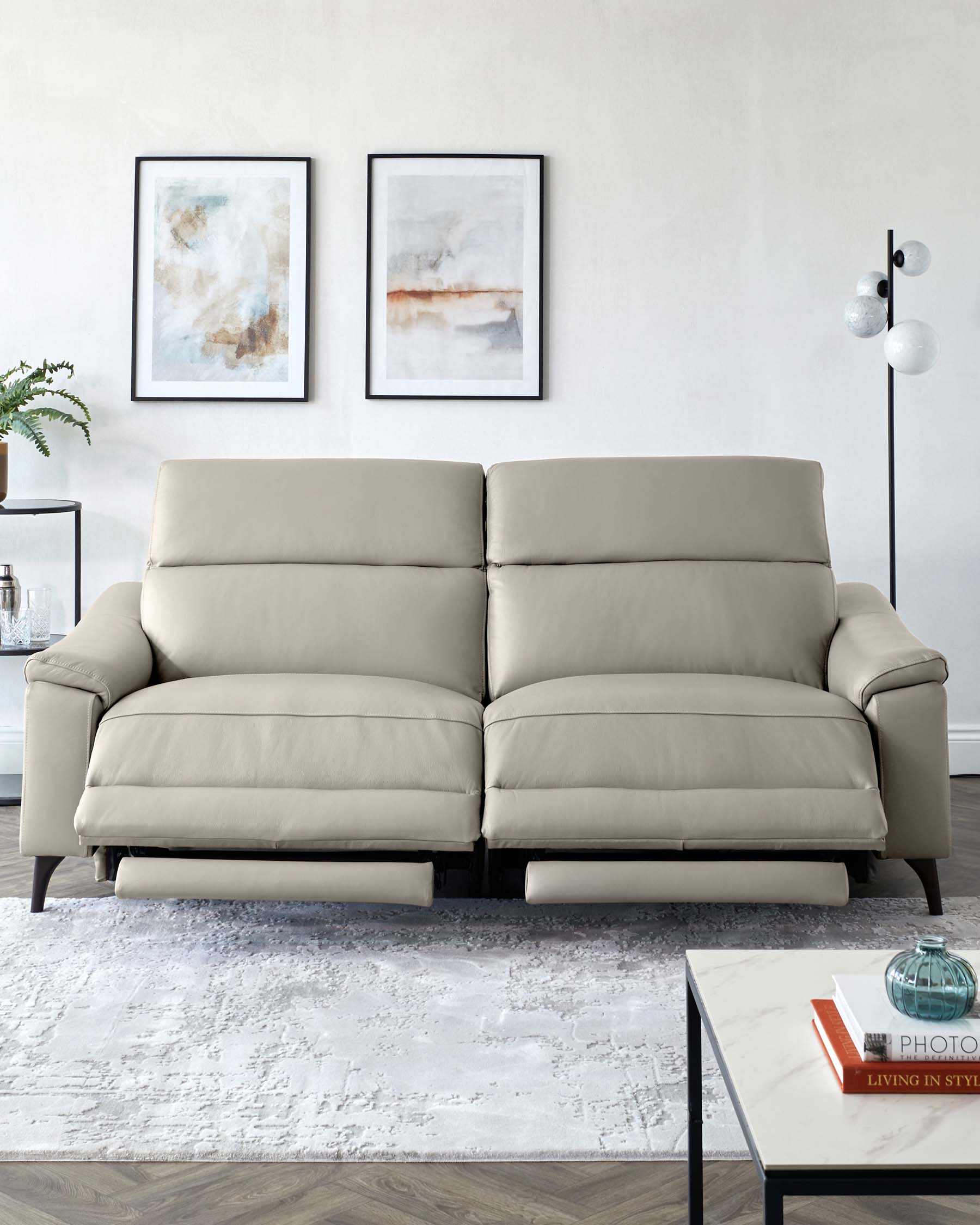 Modern neutral-toned leather sofa with plush cushions and dark wooden legs, complemented by a white marble top coffee table with a black metal frame, set upon a textured white area rug. A sleek black floor lamp with a unique globular design adds a contemporary touch, and the ensemble is balanced by two abstract framed artworks above the sofa.