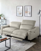 A contemporary light grey leather reclining sofa with plush back and seat cushions, flanked by a minimalist black-framed coffee table with a glass top, and a tiered round side table with glass shelves. The scene is accented by a textured off-white area rug, abstract wall art, and a modern floor lamp with spherical shades.