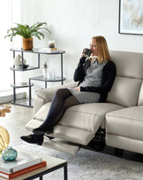 Modern living room setup featuring a plush, light beige leather sofa with recliner function, a sleek, black tiered round shelving unit housing various decorative items, and a contemporary glass-topped coffee table with a black frame, supporting books and an artistic glass vase.