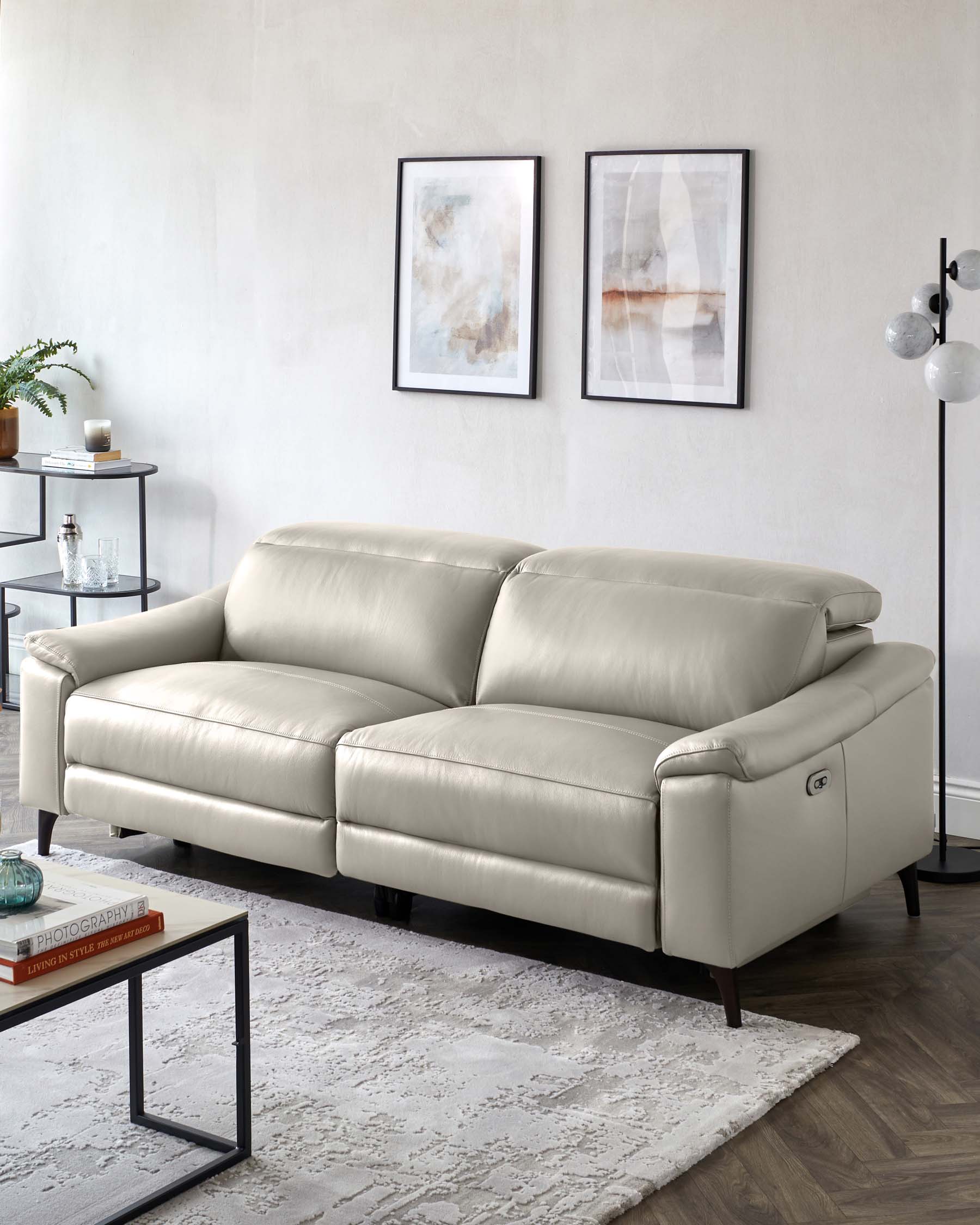 Elegant contemporary leather sofa with plush cushioning and a sleek silhouette, featuring a light taupe upholstery and subtly curved armrests, set upon dark wooden legs. Positioned in a well-lit room with modern decor, including a minimalist metal-framed side table and a neutral-toned area rug beneath.