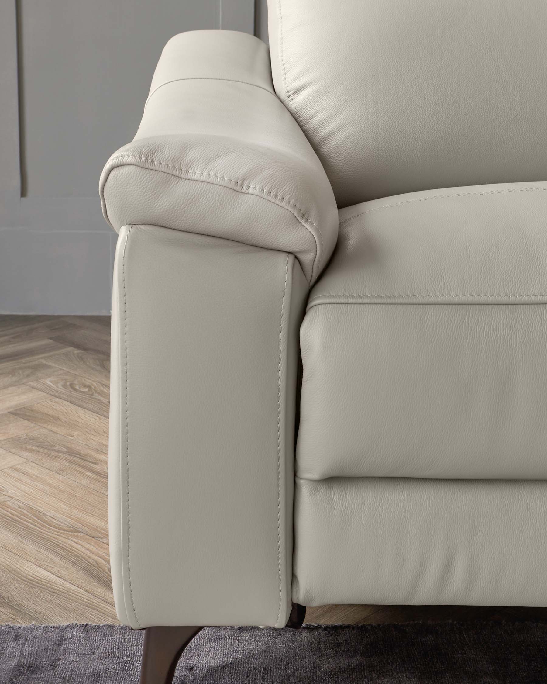 A section of a modern light beige leather sofa showing detailed stitching on its plush armrest, with a soft padded backrest and a cushioned seat mounted on a tapered dark wooden leg. Visible against a backdrop featuring grey walls and wooden flooring.