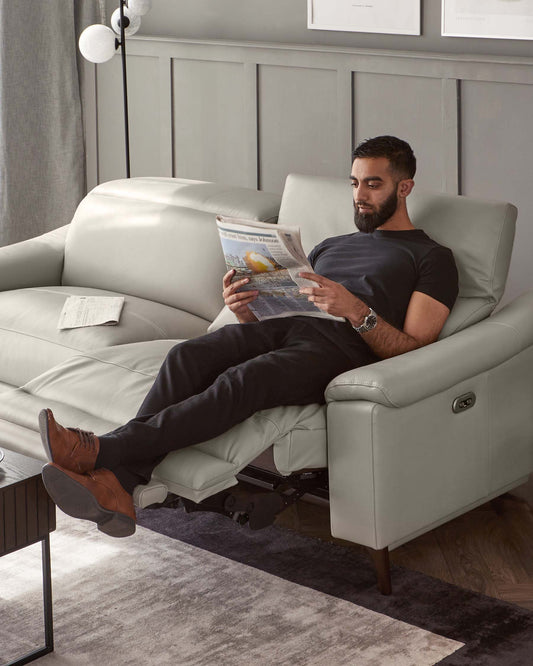 A modern light grey leather recliner sofa with plush cushioning and an extended footrest. The sofa features a sleek design with simple, clean lines, slightly rounded armrests, and dark wooden legs. There is a small black rectangular side table with a minimalistic style next to the sofa.