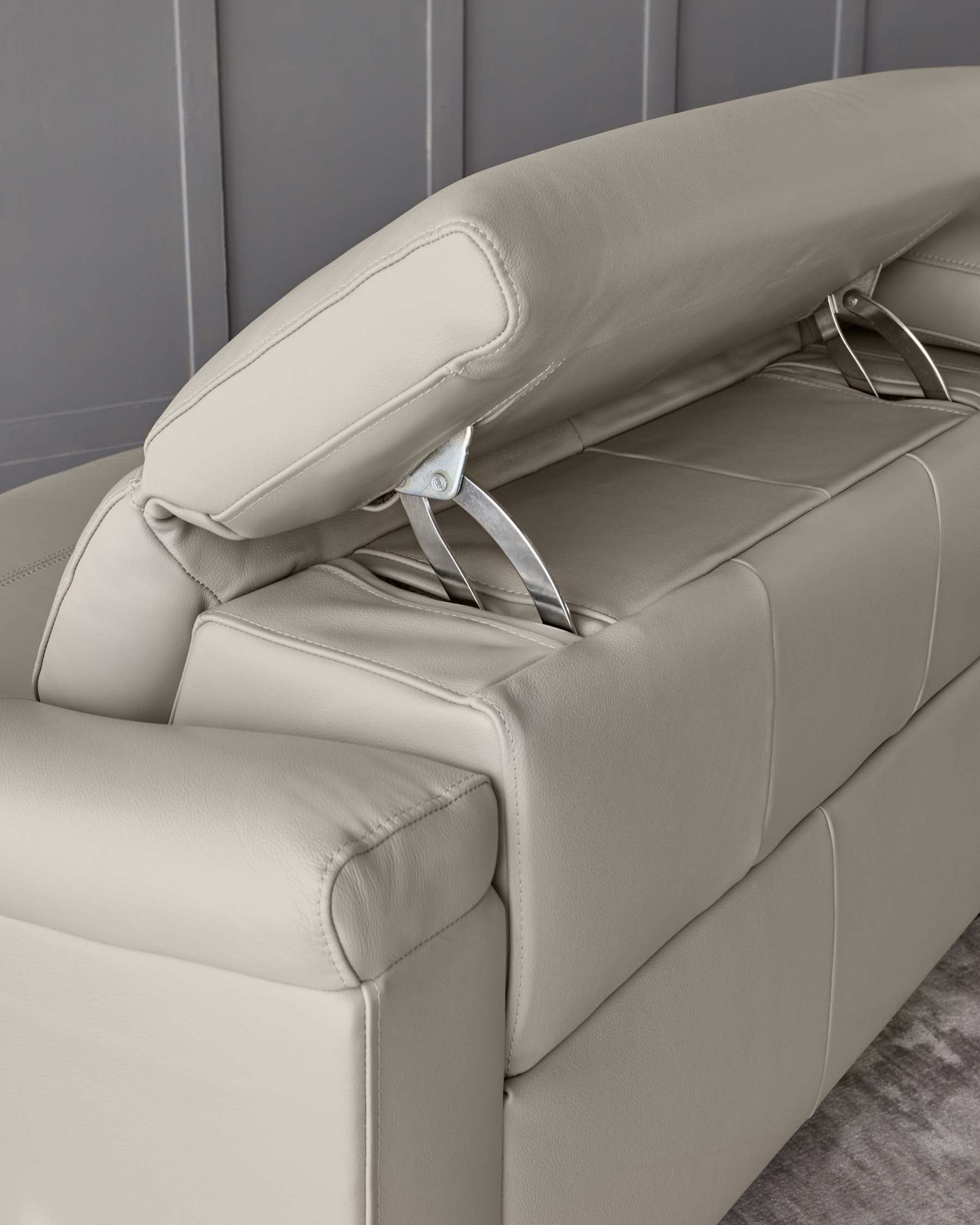 A modern light grey leather recliner sofa featuring a plush padded headrest, detailed stitching, and a manual pull lever on the side with metallic accents. The sofa is set against a grey panelled wall, highlighting its sleek and contemporary design.