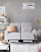 A contemporary light grey fabric two-seater sofa with reclining footrests and flanked arms, complemented by a pale pink cushion. The sofa is situated on a faded beige and white patterned area rug, with a white side table to the left and decorative elements throughout the room, including a spherical textured vase with dried floral stems on a white credenza, a framed abstract art piece, and a striking modern pendant light with vertical elements above.