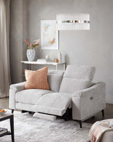 A contemporary light grey fabric reclining sofa with plush back cushions, padded armrests, and an extended footrest on one side. A pastel pink decorative pillow complements the couch, situated on a textured white area rug. In the background, a clean-lined white console table holds decorative vases and books beneath a large framed abstract artwork, adjacent to an elegant pendant light with vertical fringe details.