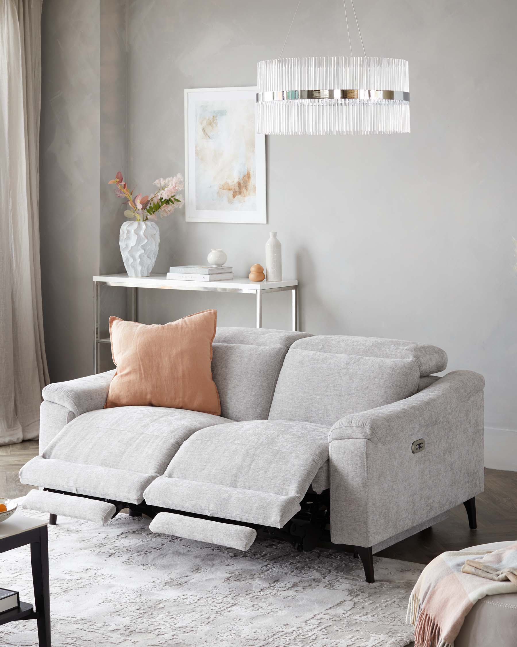 Contemporary light grey fabric reclining sofa with tufted back and extendable footrests, paired with peach accent pillow, on a muted abstract area rug. In the background, a simple white console table with decorative vases and books under an elegant hanging chandelier.