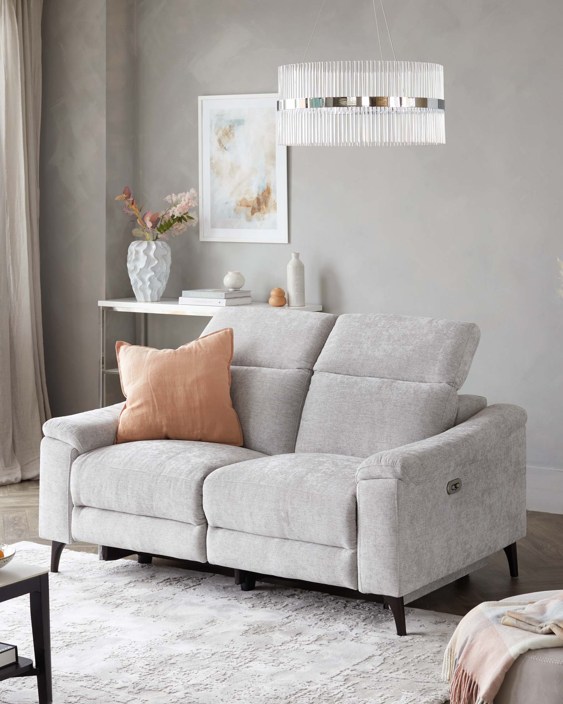 A modern light grey fabric loveseat with plush cushioning, featuring adjustable headrests, simple arm design, and dark wooden legs. The sofa is accessorized with a soft pink throw pillow, and sits on a textured white area rug. A sleek white console table with decorative vases, books, and a chic hanging pendant light with a fringe design are visible in the background.